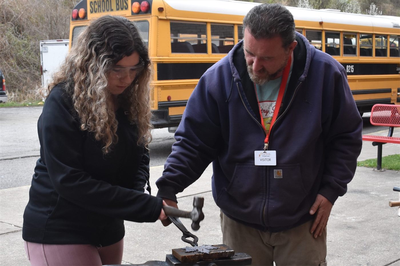 A student and a blacksmith hammer metal on an anvil