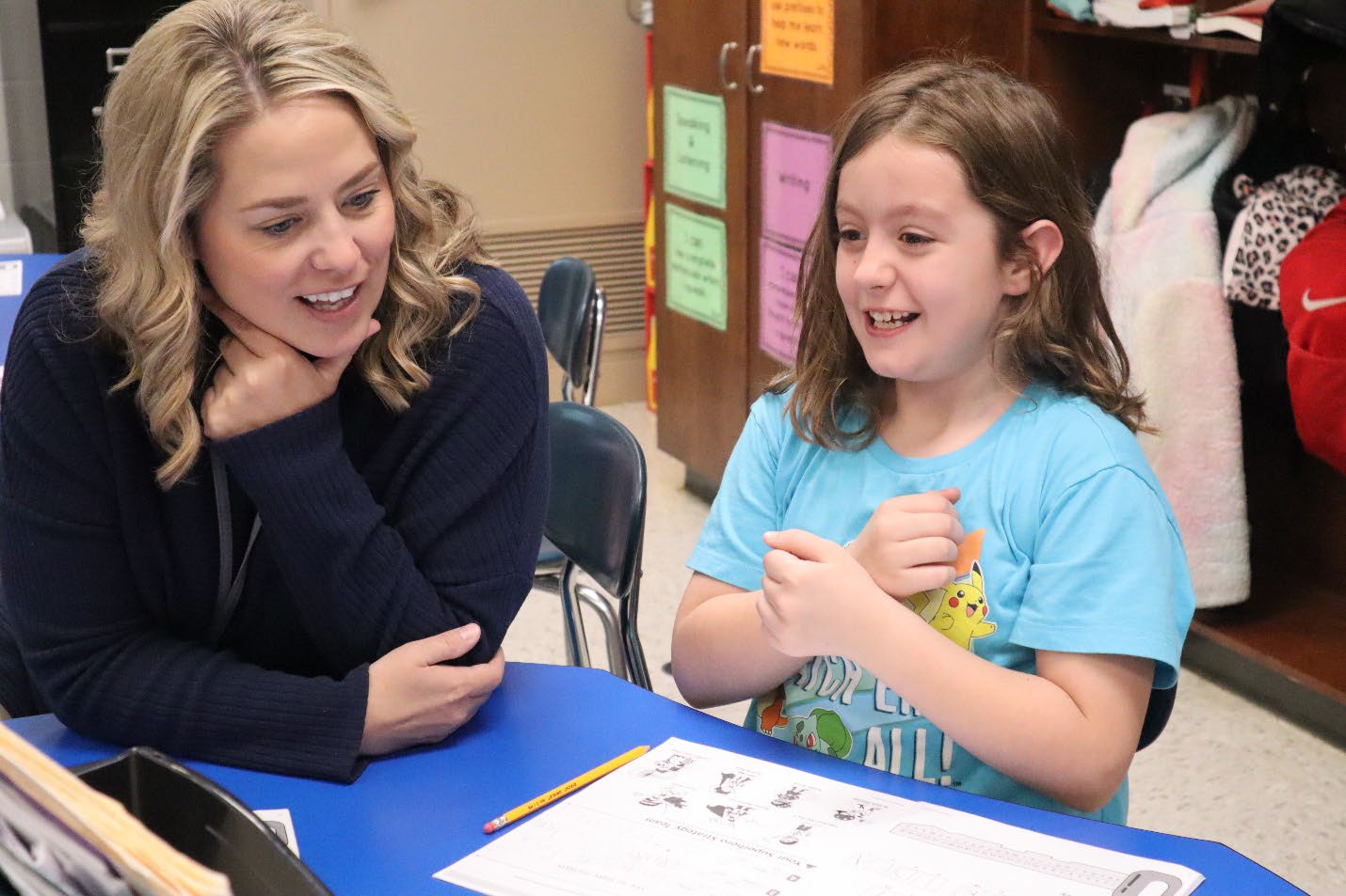 A smiling woman sits next to a smiling student at a desk